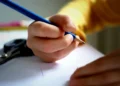 Mano di un ragazzo scrive con una matita blu su un foglio bianco, sullo sfondo forbici e pastelli colorati sfocati