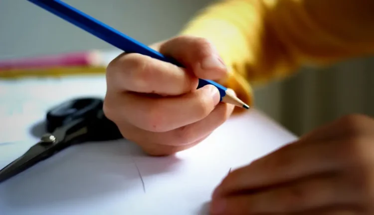Mano di un ragazzo scrive con una matita blu su un foglio bianco, sullo sfondo forbici e pastelli colorati sfocati