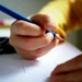 Mano di un ragazzo scrive con una matita blu su un foglio bianco, sullo sfondo forbici e pastelli colorati sfocati