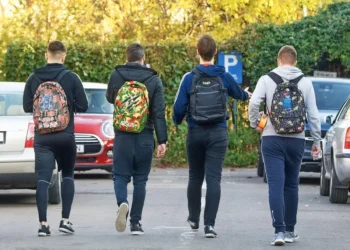 Quattro studenti di spalle con zaini colorati camminano in un parcheggio alberato, in una giornata autunnale