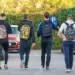 Quattro studenti di spalle con zaini colorati camminano in un parcheggio alberato, in una giornata autunnale