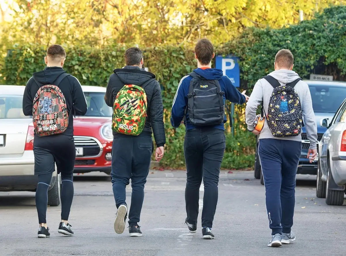 Quattro studenti di spalle con zaini colorati camminano in un parcheggio alberato, in una giornata autunnale