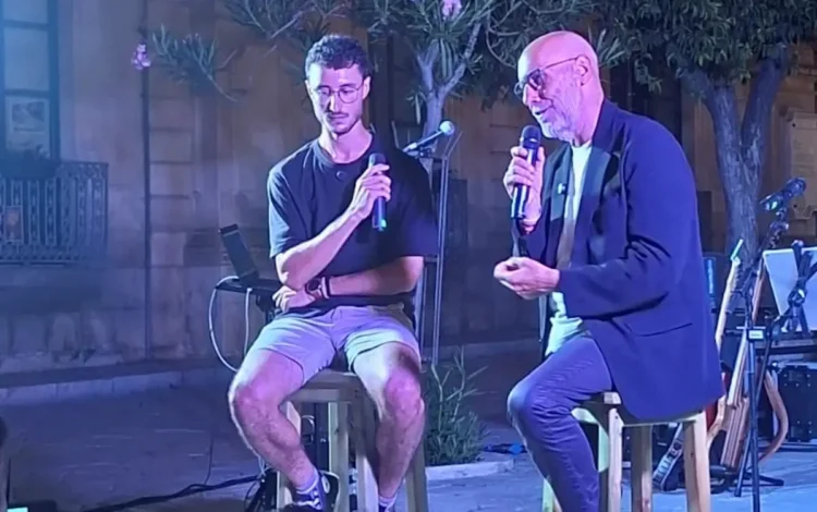Due uomini seduti su sgabelli con microfono durante una serata pubblica davanti a un edificio in pietra con balcone.