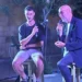 Due uomini seduti su sgabelli con microfono durante una serata pubblica davanti a un edificio in pietra con balcone.