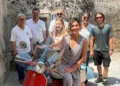 Gloria Bortolini accanto a una Vespa d'epoca rossa e celeste, con i soci del Vespa Club Modica in un vicolo di Modica.
