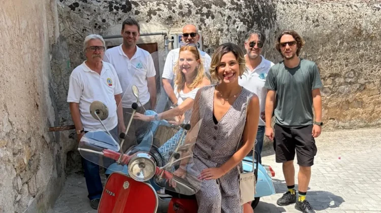 Gloria Bortolini accanto a una Vespa d'epoca rossa e celeste, con i soci del Vespa Club Modica in un vicolo di Modica.