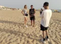 Un'inviata Rai e un istruttore con racchette da beach tennis in spiaggia a Scoglitti, ripresi da un operatore video