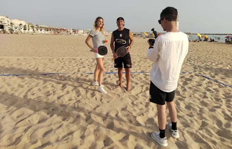Un'inviata Rai e un istruttore con racchette da beach tennis in spiaggia a Scoglitti, ripresi da un operatore video