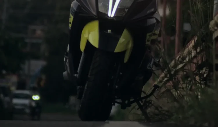 Scooter nero e giallo ripreso di notte su una strada urbana, con vegetazione sul lato destro e un'auto sullo sfondo