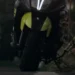 Scooter nero e giallo ripreso di notte su una strada urbana, con vegetazione sul lato destro e un'auto sullo sfondo