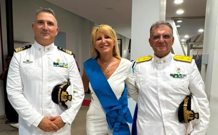 Una donna con fascia azzurra cerimoniale affiancata da due ufficiali in uniforme bianca della Guardia Costiera