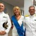 Una donna con fascia azzurra cerimoniale affiancata da due ufficiali in uniforme bianca della Guardia Costiera