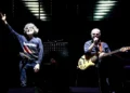 Due musicisti sul palco: uno con la mano alzata, l'altro al basso e al microfono, in un concerto della PFM