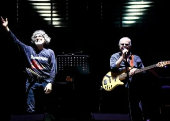 Due musicisti sul palco: uno con la mano alzata, l'altro al basso e al microfono, in un concerto della PFM