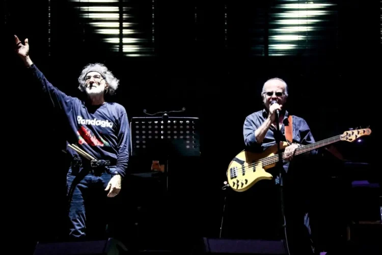 Due musicisti sul palco: uno con la mano alzata, l'altro al basso e al microfono, in un concerto della PFM