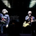 Due musicisti sul palco: uno con la mano alzata, l'altro al basso e al microfono, in un concerto della PFM