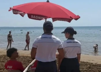 Due agenti della Guardia Costiera in divisa bianca sorvegliano una spiaggia affollata sotto un ombrellone rosso