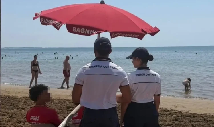 Due agenti della Guardia Costiera in divisa bianca sorvegliano una spiaggia affollata sotto un ombrellone rosso