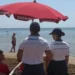 Due agenti della Guardia Costiera in divisa bianca sorvegliano una spiaggia affollata sotto un ombrellone rosso