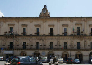 Palazzo comunale di Modica in stile barocco, con orologio in facciata, bandiera italiana e auto parcheggiate in piazza.