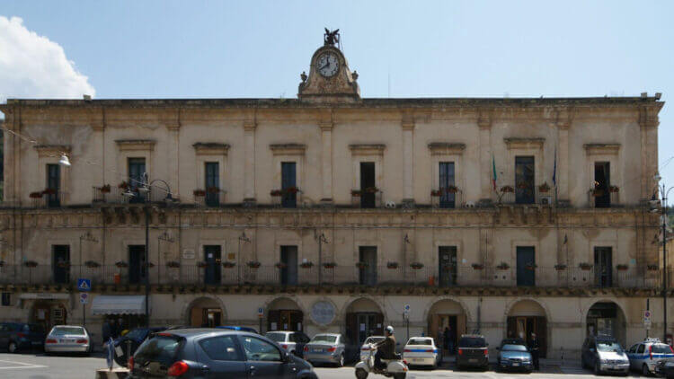 Palazzo comunale di Modica in stile barocco, con orologio in facciata, bandiera italiana e auto parcheggiate in piazza.