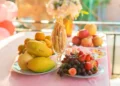 Tavolo imbandito con vassoi di frutta fresca di stagione — uva, pere, mandarini, fragole e mele — tra fiori e decorazioni