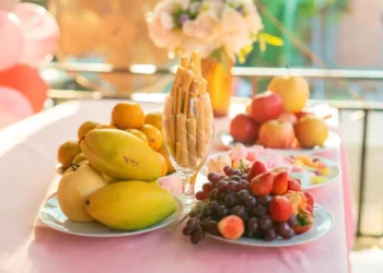 Tavolo imbandito con vassoi di frutta fresca di stagione — uva, pere, mandarini, fragole e mele — tra fiori e decorazioni