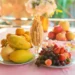 Tavolo imbandito con vassoi di frutta fresca di stagione — uva, pere, mandarini, fragole e mele — tra fiori e decorazioni