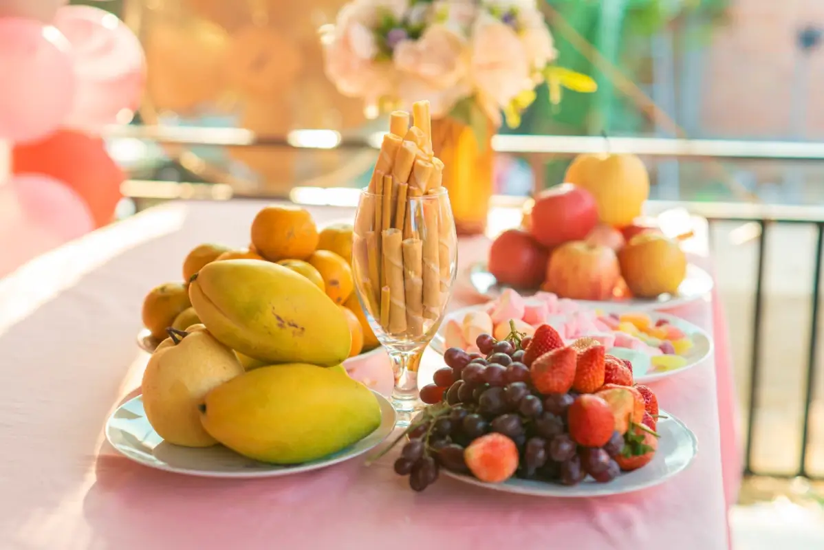 Tavolo imbandito con vassoi di frutta fresca di stagione — uva, pere, mandarini, fragole e mele — tra fiori e decorazioni