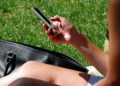 Una persona seduta sull'erba scorre lo schermo dello smartphone tenuto in mano, con una borsa nera accanto