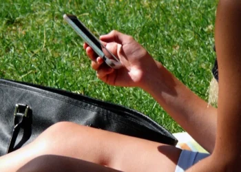 Una persona seduta sull'erba scorre lo schermo dello smartphone tenuto in mano, con una borsa nera accanto