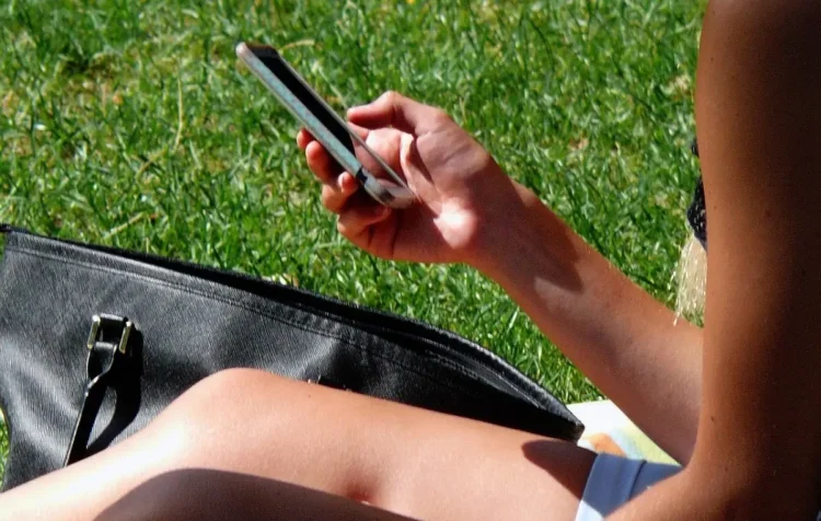 Una persona seduta sull'erba scorre lo schermo dello smartphone tenuto in mano, con una borsa nera accanto
