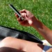 Una persona seduta sull'erba scorre lo schermo dello smartphone tenuto in mano, con una borsa nera accanto