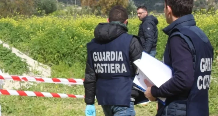 Agenti della Guardia Costiera in giubbotto operativo ispezionano un'area recintata con nastro bianco-rosso in aperta campagna