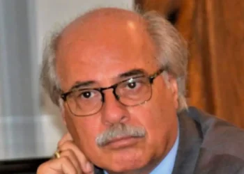 Ritratto di un uomo anziano con baffi e occhiali, giacca grigia, mano al mento, sguardo verso l'obiettivo.
