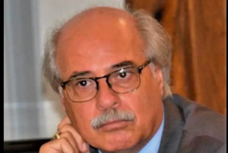 Ritratto di un uomo anziano con baffi e occhiali, giacca grigia, mano al mento, sguardo verso l'obiettivo.