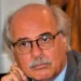 Ritratto di un uomo anziano con baffi e occhiali, giacca grigia, mano al mento, sguardo verso l'obiettivo.