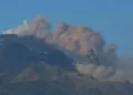 Colonna di cenere grigia si alza dal cratere dell'Etna su cielo sereno, con neve residua sul versante