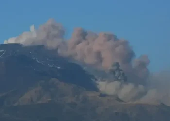 Colonna di cenere grigia si alza dal cratere dell'Etna su cielo sereno, con neve residua sul versante