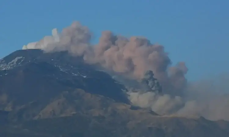 Colonna di cenere grigia si alza dal cratere dell'Etna su cielo sereno, con neve residua sul versante