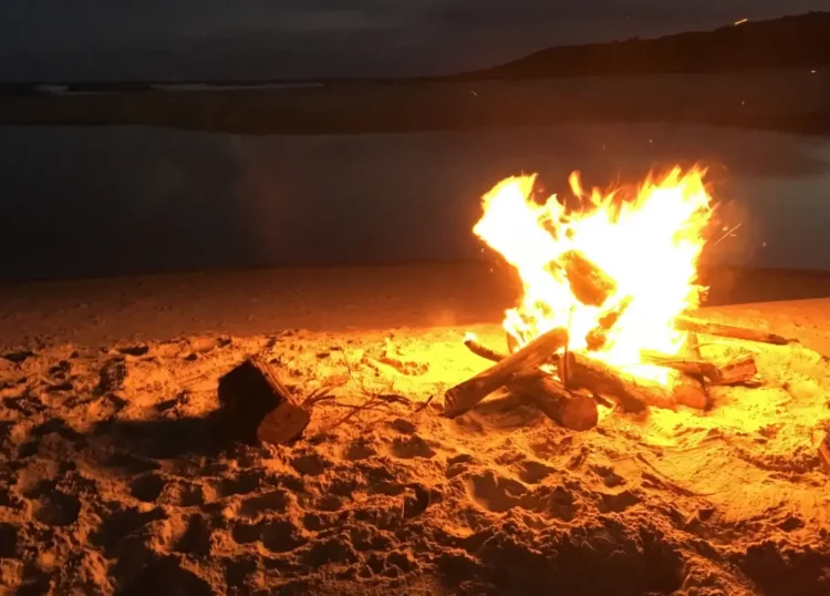 Falò acceso di notte su una spiaggia sabbiosa, con fiamme alte e braci attorno a tronchi di legno