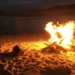 Falò acceso di notte su una spiaggia sabbiosa, con fiamme alte e braci attorno a tronchi di legno