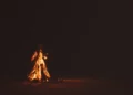 Falò acceso di notte su una spiaggia sabbiosa, fiamme arancioni e scintille su sfondo scuro.