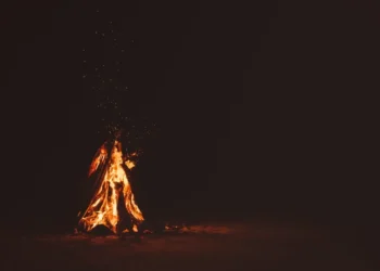 Falò acceso di notte su una spiaggia sabbiosa, fiamme arancioni e scintille su sfondo scuro.