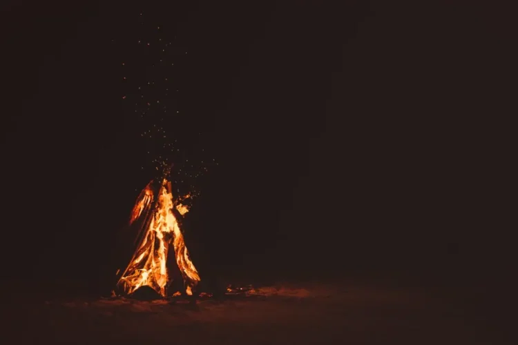 Falò acceso di notte su una spiaggia sabbiosa, fiamme arancioni e scintille su sfondo scuro.