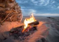 Falò acceso sulla sabbia al tramonto, con tronchi di legno tra le braci e cielo nuvoloso sullo sfondo