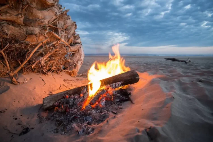 Falò acceso sulla sabbia al tramonto, con tronchi di legno tra le braci e cielo nuvoloso sullo sfondo