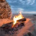 Falò acceso sulla sabbia al tramonto, con tronchi di legno tra le braci e cielo nuvoloso sullo sfondo