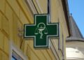 Insegna verde di una farmacia con il simbolo del mortaio e del serpente, su una parete gialla d'angolo.