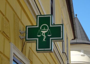 Insegna verde di una farmacia con il simbolo del mortaio e del serpente, su una parete gialla d'angolo.